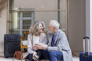 Glückliches älteres Paar mit Gepäck, das vor einem Boutique-Hotel sitzt - EIF03806