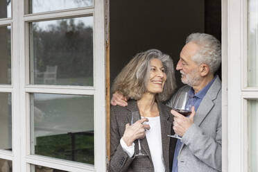 Smiling senior couple with wineglasses standing at doorway - EIF03818