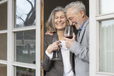 Cheerful senior couple with wineglasses standing at doorway - EIF03819