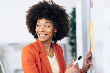 Glückliche Geschäftsfrau mit Afrofrisur, die an einem Whiteboard im Büro steht - GIOF15402