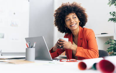 Lächelnde Geschäftsfrau mit Kaffeetasse und Laptop am Schreibtisch im Büro - GIOF15413