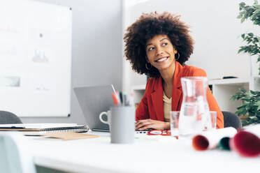 Lächelnde Geschäftsfrau mit Laptop sitzt am Schreibtisch im Büro - GIOF15414