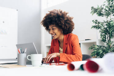 Happy businesswoman with Afro hairstyle using laptop at desk in office - GIOF15416