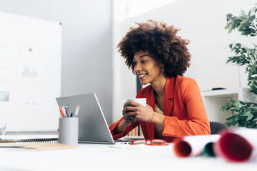 Smiling businesswoman holding coffee cup looking at laptop sitting in office - GIOF15417