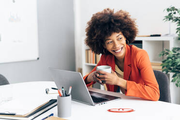 Cheerful businesswoman holding coffee cup sitting with laptop at desk in office - GIOF15418
