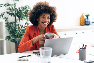 Smiling businesswoman holding coffee cup sitting with laptop at desk - GIOF15419