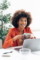 Glückliche Geschäftsfrau mit Kaffeetasse und Laptop am Schreibtisch sitzend - GIOF15420