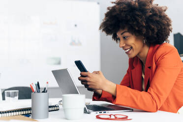 Glückliche Geschäftsfrau, die ein Smartphone benutzt und mit einem Laptop am Schreibtisch im Büro sitzt - GIOF15421