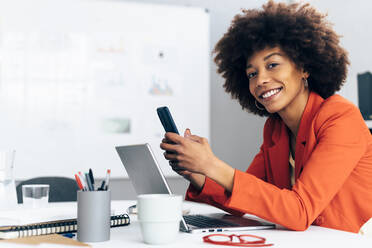 Lächelnde Geschäftsfrau mit Handy in der Hand und Laptop am Schreibtisch - GIOF15422