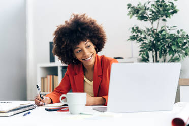 Lächelnde Geschäftsfrau mit Afrofrisur, die in ein Tagebuch schreibt und auf einen Laptop im Büro schaut - GIOF15426