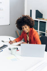 Smiling businesswoman writing in diary looking at laptop in office - GIOF15427