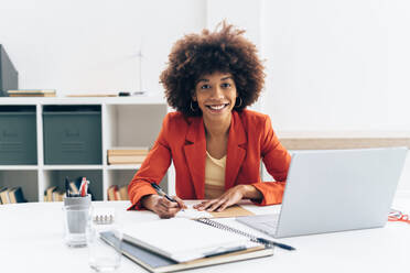Lächelnde Geschäftsfrau mit Afrofrisur sitzt mit Laptop am Schreibtisch im Büro - GIOF15429