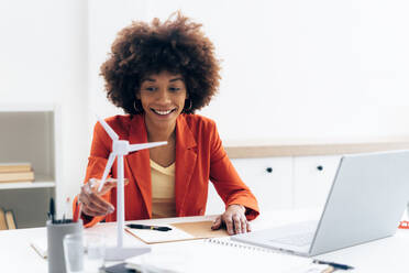 Smiling businesswoman with windmill model and laptop sitting at desk in office - GIOF15431