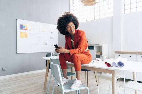 Smiling businesswoman with mobile phone sitting on desk in office - GIOF15437