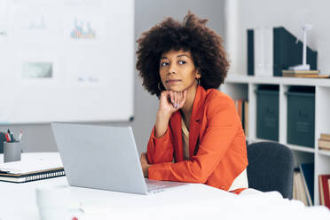 Nachdenkliche Geschäftsfrau mit Hand am Kinn sitzt mit Laptop am Schreibtisch im Büro - GIOF15449