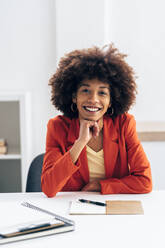 Smiling businesswoman with hand on chin sitting at desk in office - GIOF15450