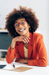 Cheerful businesswoman wearing eyeglasses sitting with hand on chin at desk - GIOF15460