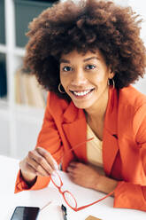 Smiling businesswoman holding eyeglasses sitting at desk - GIOF15462