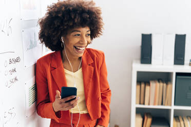 Cheerful businesswoman holding smart phone listening music through in-ear headphones at office - GIOF15465