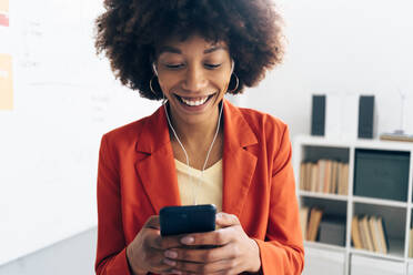 Happy businesswoman using smart phone listening music through in-ear headphones at office - GIOF15468