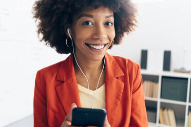 Lächelnde Geschäftsfrau mit Afrofrisur hört Musik über In-Ear-Kopfhörer im Büro - GIOF15470