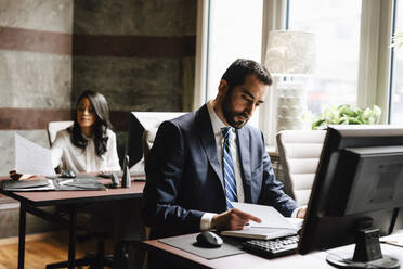 Confident male lawyer reading diary with female colleague working in background at office - MASF29278