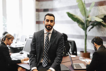 Portrait of bearded businessman sitting on desk in office - MASF29296