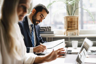 Businessman analyzing document by businesswoman using laptop at desk in office - MASF29343