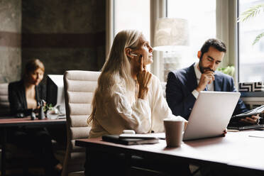 Mature businesswoman wearing in-ear headphones at laptop by businessman at desk in office - MASF29345