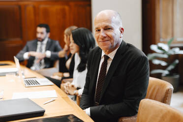 Portrait of smiling senior male lawyer in board room with colleagues during meeting - MASF29375