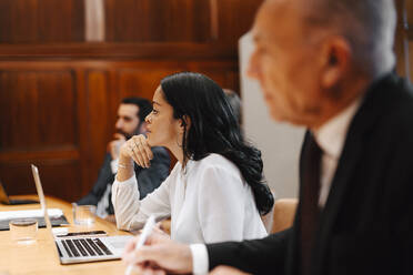 Geschäftsfrau mit Hand am Kinn sitzt inmitten männlicher Kollegen in einer Vorstandssitzung - MASF29386