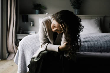 Emotionally depressed woman sitting with hand in hair against bed at home - MASF29438