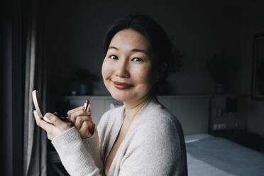 Portrait of smiling woman applying lipstick in bedroom at home - MASF29465