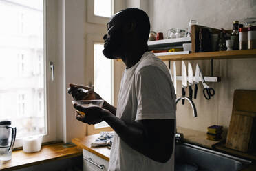 Seitenansicht eines lächelnden Mannes, der eine Schüssel und eine Gabel hält und in der Küche zu Hause isst - MASF29478