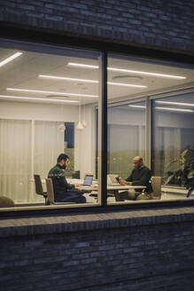 Businessman with male colleague working late in office seen through window at night - MASF29524