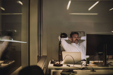 Mature businessman with hands behind head working in office at night - MASF29544