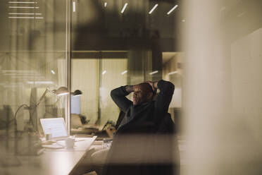 Mature businessman with hands behind head working overtime in office at night - MASF29555