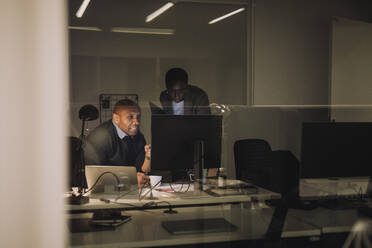 Businessman discussing with female colleague over computer seen through glass at work place - MASF29559