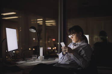 Geschäftsfrau Textnachrichten auf Smartphone sitzend am Arbeitsplatz - MASF29605
