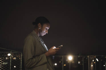 Seitenansicht einer Geschäftsfrau, die nachts eine Textnachricht auf einem Mobiltelefon schreibt - MASF29614