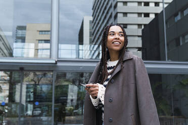 Smiling businesswoman wearing blazer holding smart phone standing at office park - PNAF03623