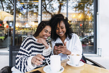 Glückliche Kollegen, die in einem Straßencafé sitzen und sich ein Smartphone teilen - PNAF03650