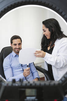 Lächelnder Patient mit Arzt, der sein Gesicht nach einer Botox-Behandlung in einer Schönheitsklinik im Spiegel untersucht - MTBF01214