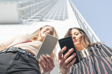 Smiling young women holding mobile phone standing in front of building - FBAF01978