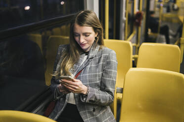 Lächelnde junge Frau, die ein Mobiltelefon benutzt, sitzt am Fenster in einer Straßenbahn - FBAF01992