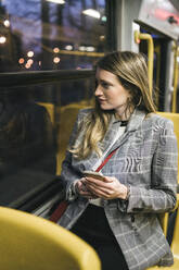 Junge Frau mit Smartphone sitzt am Fenster in der Straßenbahn - FBAF01993