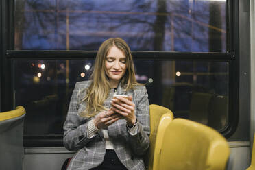 Lächelnde Frau mit Smartphone in der Straßenbahn sitzend - FBAF01995