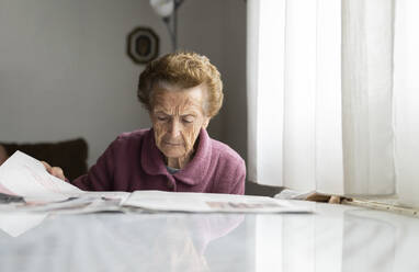 Senior woman reading newspaper at dining table - JCCMF06017