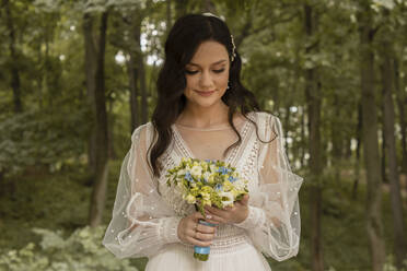 Smiling bride holding bouquet standing in front of tress - SSGF00662
