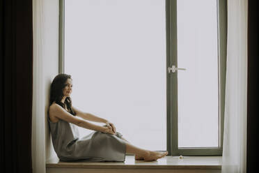 Smiling young woman sitting on window at home - SSGF00670